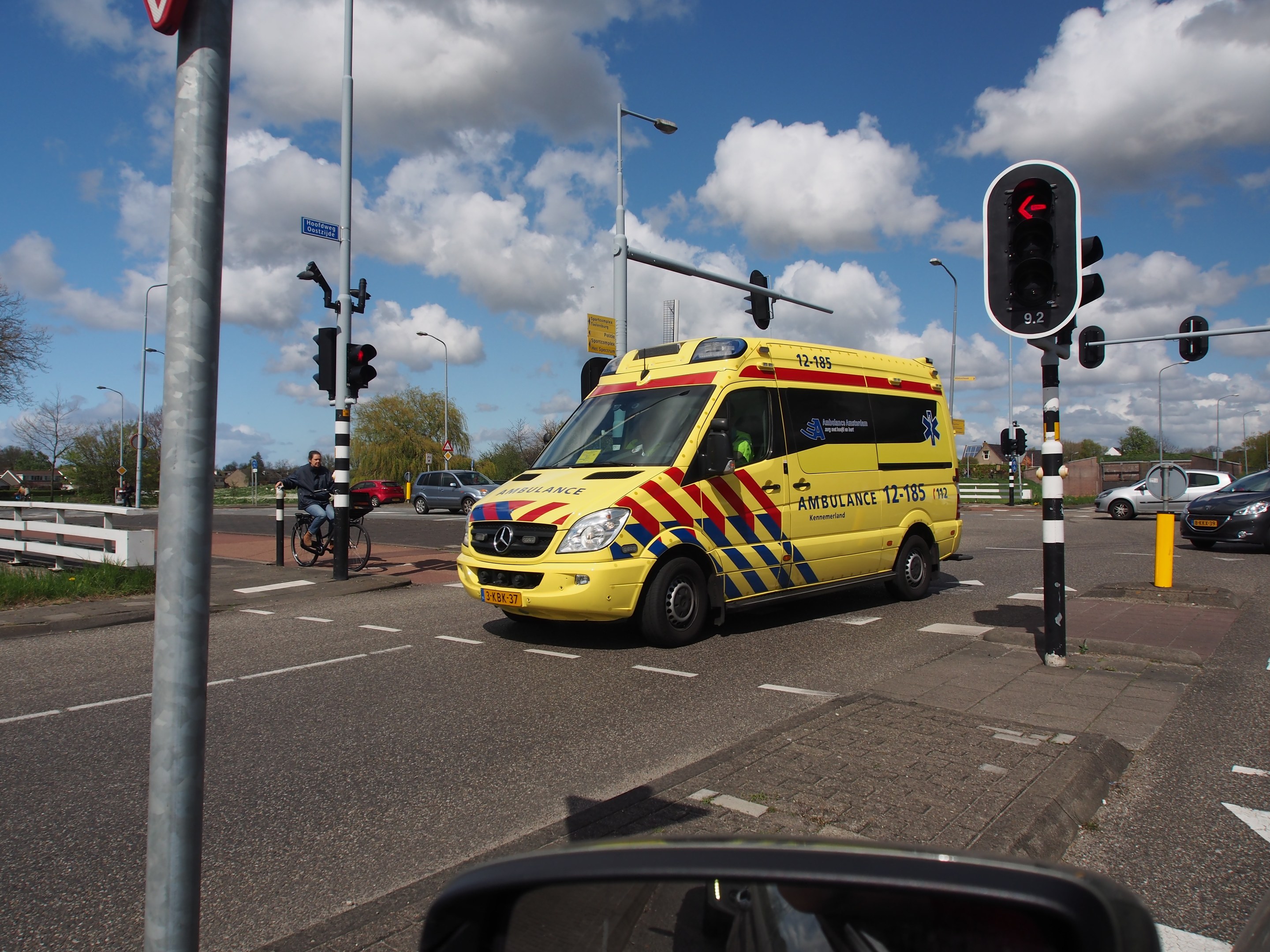 Ambulanz fährt auf einer Straße neben einer Ampel, mit einem Radfahrer im Vordergrund und Gebäuden, Bäumen, Laternen und einem bewölkten Himmel im Hintergrund.