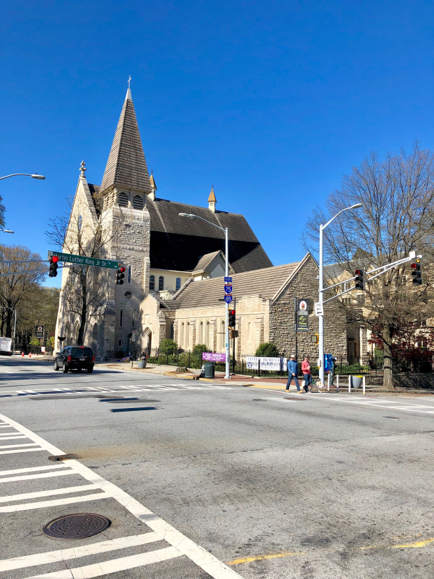 Eine große Kirche mit einem Turm ist von Gebäuden, Straßenlaternen, Verkehrszeichen, Kraftfahrzeugen, Fußgängern und Bäumen unter einem klaren blauen Himmel umgeben.