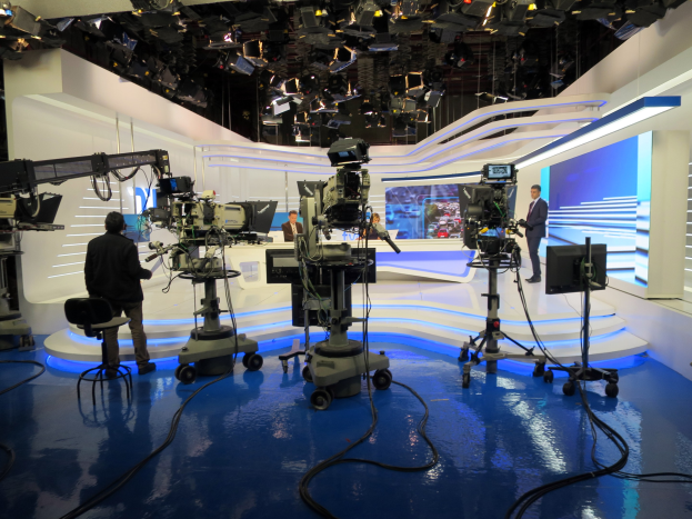 Ein TV-Studio mit mehreren bodengestützten Kameras, einer sitzenden Person im Vordergrund, einigen stehenden Personen im Hintergrund und deckengestützten Lampen, die auf eine Live-Übertragung hinweisen.