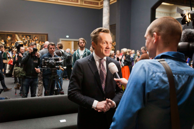 Ein Mann im Anzug und Krawatte spricht mit einem Reporter vor einer Menge, einige halten Kameras, mit einem Sofa im Vordergrund und einer Wand mit einem Foto, Lampen und einer Säule im Hintergrund, während er ein Mikrofon hält.