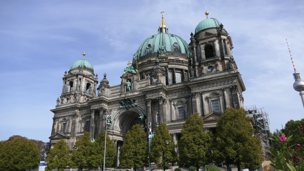 Berliner Dom, eine große Kathedrale mit Säulen, Bögen, Statuen und Fenstern, vor einem bewölkten Himmel, mit Menschen, Bäumen und einem Turm im Vordergrund.