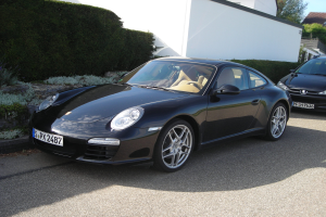 Zwei schwarze Porsche 911 vor einem weißen Haus geparkt, umgeben von üppiger Vegetation und Bäumen, mit einem hellblauen Himmel im Hintergrund.
