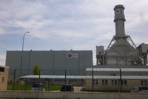 Ein großes Gebäude mit einem zentralen Turm, umgeben von einem Zaun, mit Fahrzeugen auf der benachbarten Straße, Polen, Lampen, Drähten, Bäumen und Gras im Vordergrund und einem bewölkten Himmel im Hintergrund, das eine neue Energiespeicheranlage darstellt.