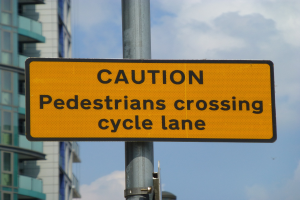Gelbes Warnschild an einem Pfahl vor einem Gebäude, mit einem klaren blauen Himmel im Hintergrund, das "Fußgänger überqueren Fahrradspur" anzeigt.