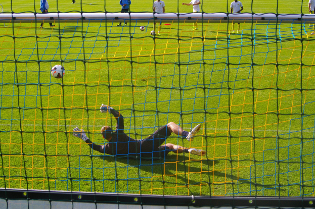 Ein Fussballspieler taucht ab, um einen Ball vor einem Tor zu halten, mit einem Netz im Vordergrund und einigen Menschen im Hintergrund, während mehrere Fussbälle auf dem Boden verstreut sind.