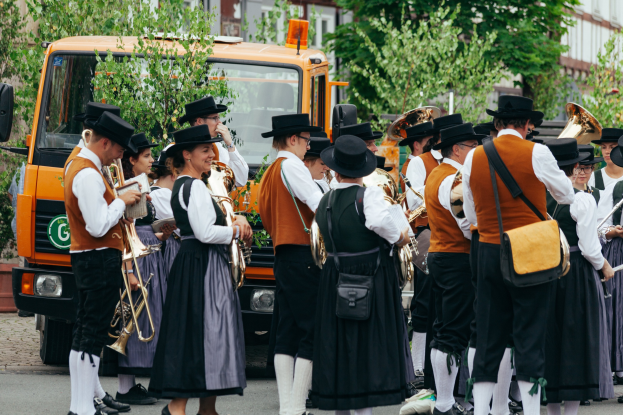 Gruppe von Menschen in traditioneller bayrischer Tracht, die Instrumente spielt und Taschen trägt, mit Bäumen und Gebäuden im Hintergrund und einem geparkten Fahrzeug.
