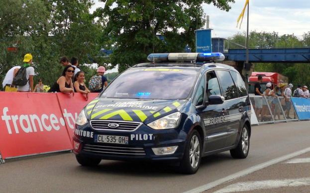 Ein Polizeiwagen fährt auf einer Straße an einer Menge von Menschen vorbei, einige tragen Mützen und Taschen, mit einem Banner links, Geländern mit Bannern dahinter und einem Hintergrund aus Bäumen, einer Brücke, einer Fahne auf einem Mast und einem bewölktem Himmel.