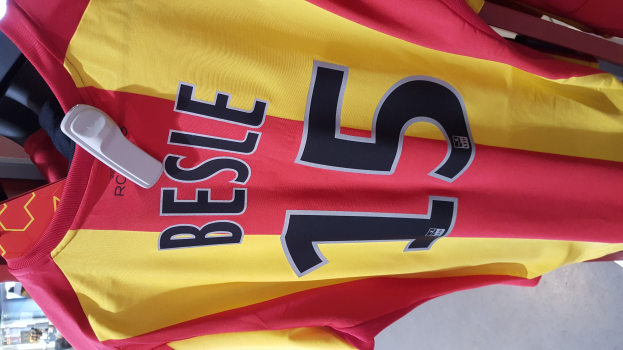 Ein rotes und gelbes Fußballtrikot mit Text und Nummern hängt an einem Ladengestell, mit anderen Gegenständen im Hintergrund.