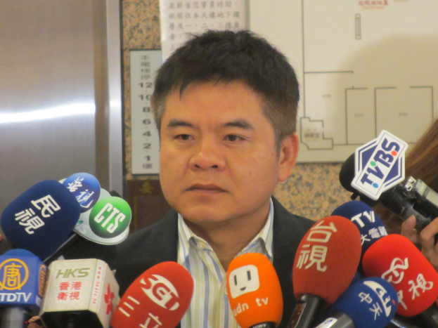 Ein Mann im Anzug und Krawatte spricht zu Reportern vor einer Wand mit einer Tafel, während eine Frau mit einer Kamera rechts steht.