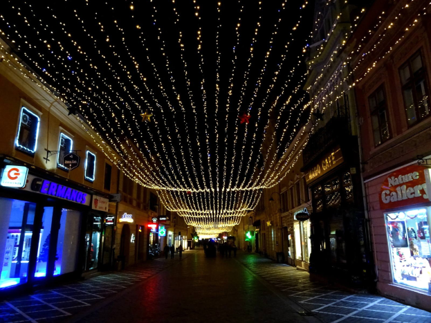 Eine nächtliche Stadtstraße mit Weihnachtsbeleuchtung, gesäumt von Gebäuden mit beleuchteten Fenstern und Schildern, mit ein paar Passanten auf dem Weg.