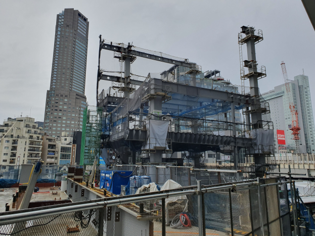 GroÃer Baustelle fÃr ein neues Kraftwerk in einer Stadtumgebung, umgeben von einem Metallzaun, mit GebÃuden, Masten, einem Banner, Fahrzeugen, DrÃhten und einem bewÃlktem Himmel.
