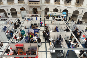 Große Gruppe von Menschen auf einer internationalen Fachmesse mit Ständen, Bannern, Tischen, Stühlen und Ballons in einem Raum mit architektonischen Bögen, Säulen, Geländern und einem Bildschirm im Hintergrund.