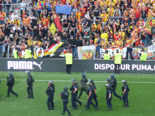 Eine Gruppe von Polizisten in Helmen und mit Waffen in der Hand geht über ein Fußballfeld, im Hintergrund halten Menschen Fahnen und Schilder nahe Absperrungen.