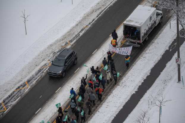 Eine Gruppe von Menschen geht eine schneebedeckte Straße entlang, trägt Schilder und Plakate und hat einen Lastwagen am Straßenrand geparkt und ein Auto fährt daneben, Bäume und Strommasten säumen die Straße.