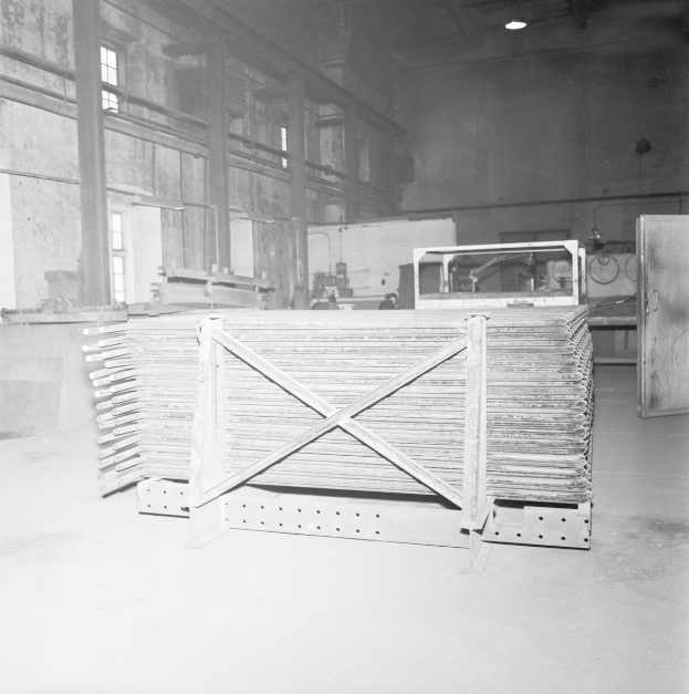 Ein Schwarz-Weiß-Foto einer Eisengießerei mit einem großen Metallgerüst, verstreuten Metallobjekten auf dem Boden, einer Tür auf der rechten Seite, einer Wand mit Fenstern im Hintergrund und Lampen oben.