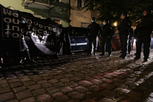 Gruppe von Polizisten in Helmen auf einem Kopfsteinpflasterweg in der Nacht stehend, ein Banner haltend, mit Fahrzeugen, Gebäuden, Bäumen und Straßenbeleuchtung im Hintergrund.
