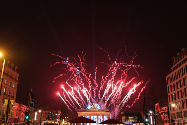 Eine belebte Stadtstraße in Berlin an Silvester, voller Menschen, Fahrzeuge und Gebäude, beleuchtet von Feuerwerk und Gebäudelichtern, die eine festliche Stimmung schaffen.
