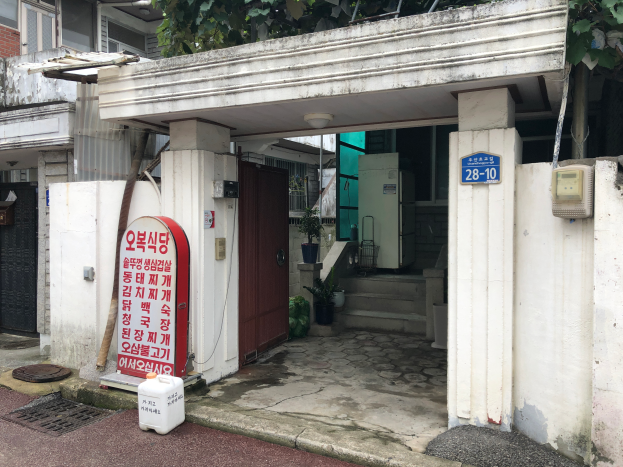 Ein Gasstation-Gebäude in Seoul, Südkorea, mit einem Schild, umgeben von Bäumen und Pflanzen, mit einer Straße unten und einer Tafel mit Text auf der linken Seite.