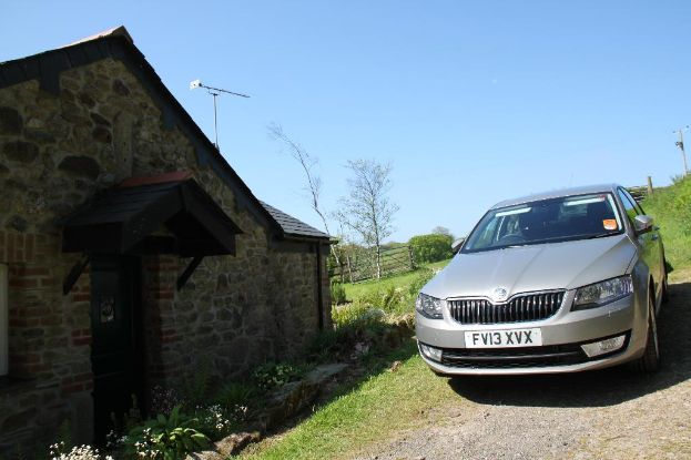 Eine Szene mit einem Haus und einem Zaun auf der linken Seite und einem Auto, Pflanzen und einem Strommast auf der rechten Seite unter einem sonnigen Himmel.