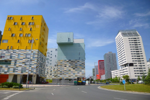 Eine Stadtstraße mit hohen Gebäuden im Hintergrund, Straßenlaternen, Autos, Informationsschildern, Büschen, Bäumen und einer bewölkten Himmel, mit einzigartig gestalteten Gebäuden der Universität Rotterdam.