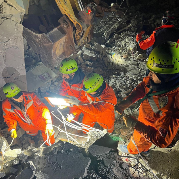 Männer in orangefarbener Uniform und grünen Helmen bei der Arbeit an Schutt mit einem Kran im Hintergrund bei einem Einsatz nach dem Einsturz eines Gebäudes.