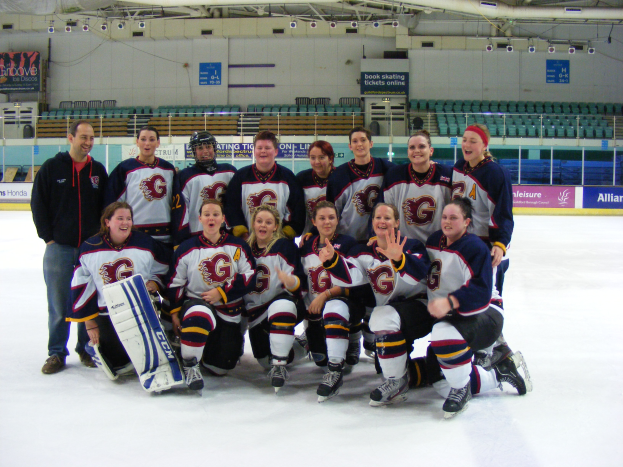 Eine Gruppe von Frauenhockeyspielerinnen in Helmen und Schlittschuhen posiert für ein Foto auf dem Eis, eine hält einen Hockeystock, mit Stühlen, Tafeln mit Text, Geländern und Deckenlampen im Hintergrund.