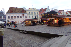 Ein geschäftiges Weihnachtsmarkt in Tallinn, Estland mit Menschen um geschmückte Stände, festliche Lichter, bewölkten Himmel, Gebäuden im Hintergrund, Treppe nach oben und Topfpflanzen.