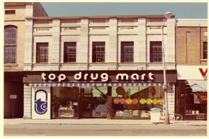 Ein Drogenmart-Geschäft an einer Straßenecke mit einem Gebäude, einem Schild, einem Straßenschild, einem geparkten Fahrrad, einem Müllcontainer und einem bewölkten Himmel.