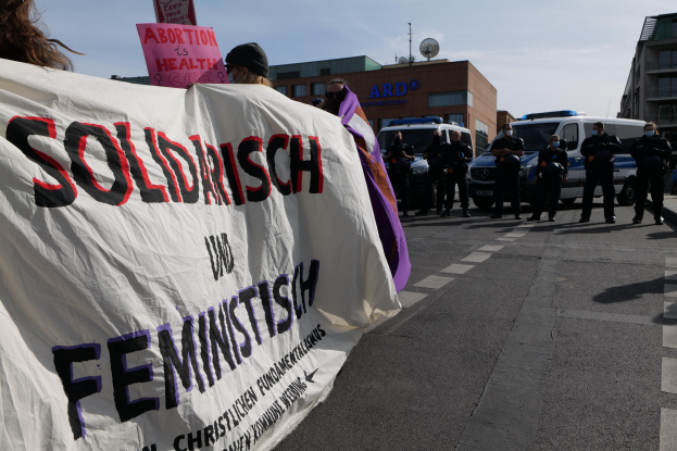 Eine Gruppe von Menschen marschiert auf einer Straße, die eine Fahne hält, auf der 'Solidarität und Feminismus' steht, mit parkenden Fahrzeugen auf der Seite und Gebäuden im Hintergrund unter einem klaren blauen Himmel.