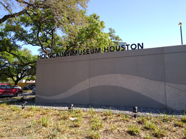 Das Holocaust-Museum in Houston, Texas, mit einer textbedeckten Wand, Steinen, Gras, Pflanzen, einer Gruppe von Bäumen, Fahrzeugen auf der Straße, einem Straßenpfahl und einem bewölkten Himmel.