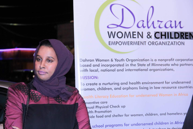 Eine Frau in einem weißen Hemd und blauen Jeans steht entschlossen vor einem Banner, auf dem "Dahran Women & Children Empowerment Organization" steht, mit einem Auto im Hintergrund.