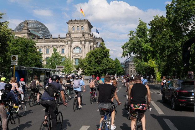 Gruppe von Menschen, die auf Fahrrädern eine von Bäumen gesäumte Straße in Berlin, Deutschland, entlangfahren, mit Gebäuden und einer Bushaltestelle im Hintergrund, unter einem bewölkten Himmel und einer Flagge an einem Gebäude.