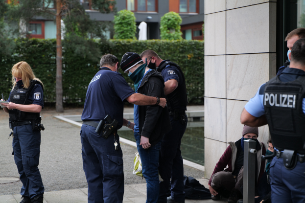 Eine Gruppe von Polizisten steht vor einem Gebäude, eine Frau hält ein Buch und einen Stift; mehrere Personen stehen und sitzen auf dem Boden, es gibt Pflanzen, Bäume und Gebäude im Hintergrund.
