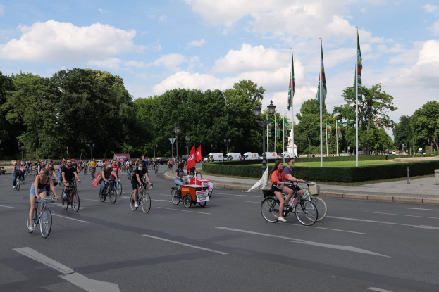 Eine Gruppe von Menschen fährt mit Fahrrädern eine von Bäumen gesäumte Straße während der Paris Bike Ride 2016 hinunter, mit Laternenmasten, Fahnenmasten, bewölktem Himmel und Fahrzeugen im Hintergrund.