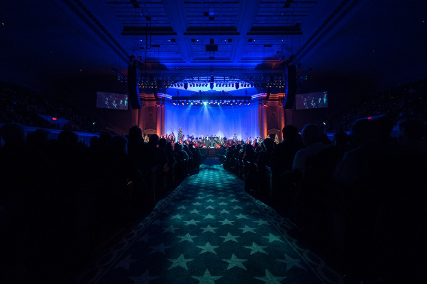 Ein großer Zuschauerraum mit sitzenden Gästen, eine Bühne mit Musikern und eine Decke mit Beleuchtung und Fernsehgeräten, was auf einen Konzertsaal während der Nationalkonvention 2019 hindeutet.