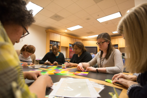 Eine Gruppe von Frauen sitzt um einen Tisch und arbeitet an einem Projekt, mit Papieren und Gegenständen auf dem Tisch, Bücherregalen im Hintergrund und Lampen an der Decke.