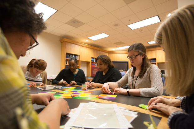 Eine Gruppe von Frauen sitzt um einen Tisch und arbeitet an einem Projekt, mit Papieren und Gegenständen auf dem Tisch, Bücherregalen im Hintergrund und Lampen an der Decke.