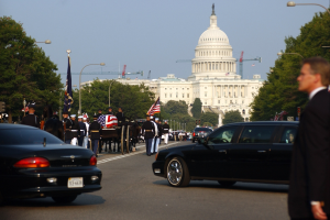 Eine Trauerprozession mit Menschen in Mützen und mit Fahnen zieht am Kapitol vorbei, flankiert von Laternenmasten, Bäumen und Gebäuden unter einem klaren Himmel.