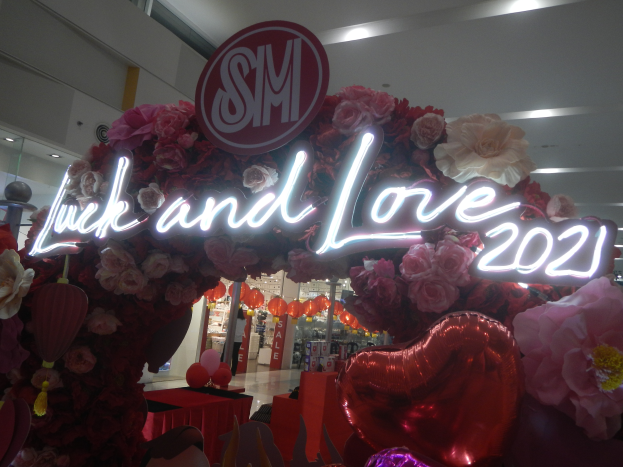 Ein Geschäft mit einem Schild "Glück und Liebe 2021" umgeben von Blumen, Ballons und anderen Dekorationen, mit anderen Geschäften, Lichtern und einer Wand im Hintergrund, die eine festliche Atmosphäre für die SM-Valentinstag-Feier schaffen.