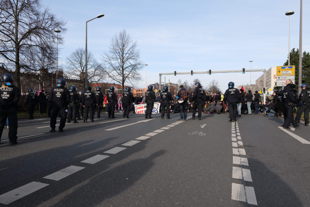 Gruppe von Polizisten in schwarzen Uniformen und Helmen, die an der Seite einer Straße mit Laternenmasten, Ampeln, Bäumen, Gebäuden und einem klaren blauen Himmel im Hintergrund stehen.