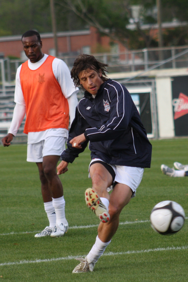 Ein Fußballspieler in einer schwarzen Jacke und weißen Shorts schießt einen Ball, während ein anderer Spieler in einer orangefarbenen Jacke hinter ihm steht und ein eingezäunter Bereich mit Bäumen im Hintergrund zu sehen ist.