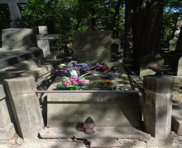 Ein Friedhof mit einem zentralen Grab, das mit Blumen und Kerzen geschmückt ist und von Bäumen im Hintergrund umgeben ist.