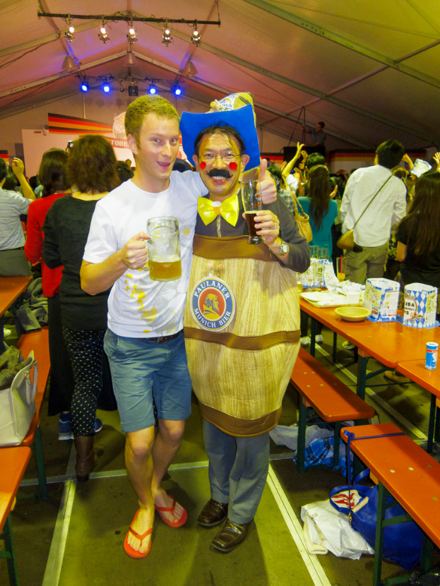 Zwei Personen stehen zusammen, eine hält ein Kind mit einem Bier, die andere trägt ein Drama-Kostüm, mit einem Tisch voller Essen daneben und einer Gruppe von Menschen im Hintergrund.