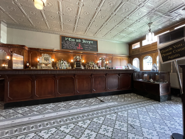 Ein großer Raum mit einer zentralen Bar, Deckenleuchten, Flaschen auf einem Tisch und einer Kreidetafel auf der rechten Seite, im Tom and Jerry Pub in London.