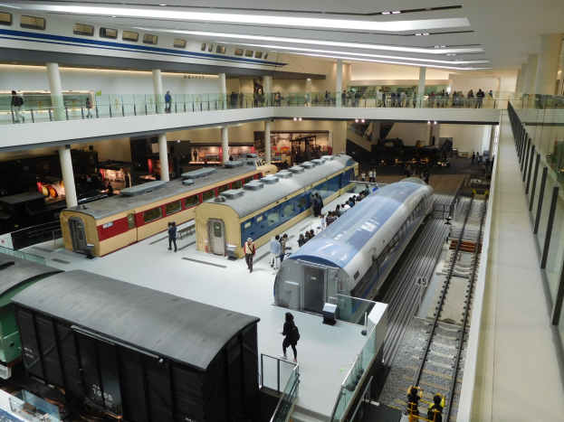 Ein großer Museumsraum mit verschiedenen Zügen in unterschiedlichen Größen und Farben, mit Menschen, die sie vom Boden, Geländern und Säulen unter Deckenbeleuchtung betrachten.