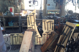 Eine Werkstatt mit verschiedenen Werkzeugen, Maschinen und Holzplanken auf Tischen, einem Stuhl links, Werkzeugen an der Wand und einem Fenster im Hintergrund.