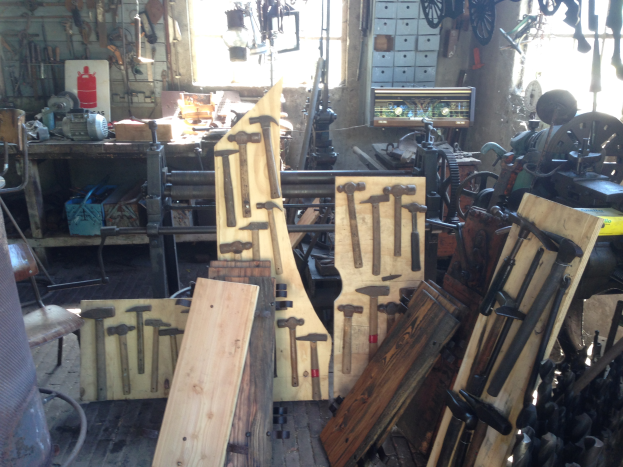 Eine Werkstatt mit verschiedenen Werkzeugen, Maschinen und Holzplanken auf Tischen, einem Stuhl links, Werkzeugen an der Wand und einem Fenster im Hintergrund.