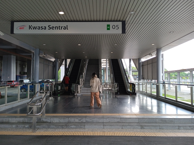 Eine Frau geht eine Rolltreppe in einem Bahnhof hinunter, mit Gelöndern, Pfeilern, Texttafeln, Deckenleuchten, Glaswänden, Straßenfahrzeugen, Bäumen und einem klaren blauen Himmel im Hintergrund.