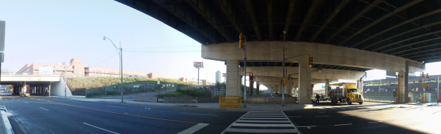 Ein Lkw fährt unter einer Brücke auf einer Stadtstraße, umgeben von Gebäuden, Straßenlaternen, Verkehrspfosten, Verkehrsampeln, Strommasten, Stromkabeln, Kraftfahrzeugen, Barrierepfosten, Büschen, Bäumen und einem klaren blauen Himmel.