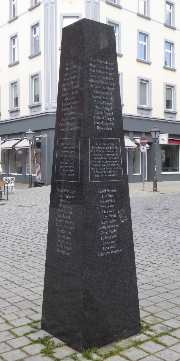 Ein hohes Denkmal mit eingravierter Inschrift steht auf einem Platz in der Stadtmitte, umgeben von Gebäuden, Straßenmöbeln und Menschen unter einem offenen Himmel.